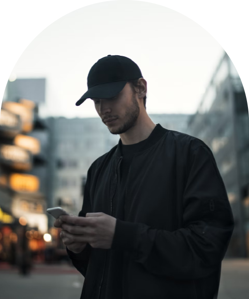 Ung man i svart keps och jacka tittar på sin telefon i en stadsmiljö